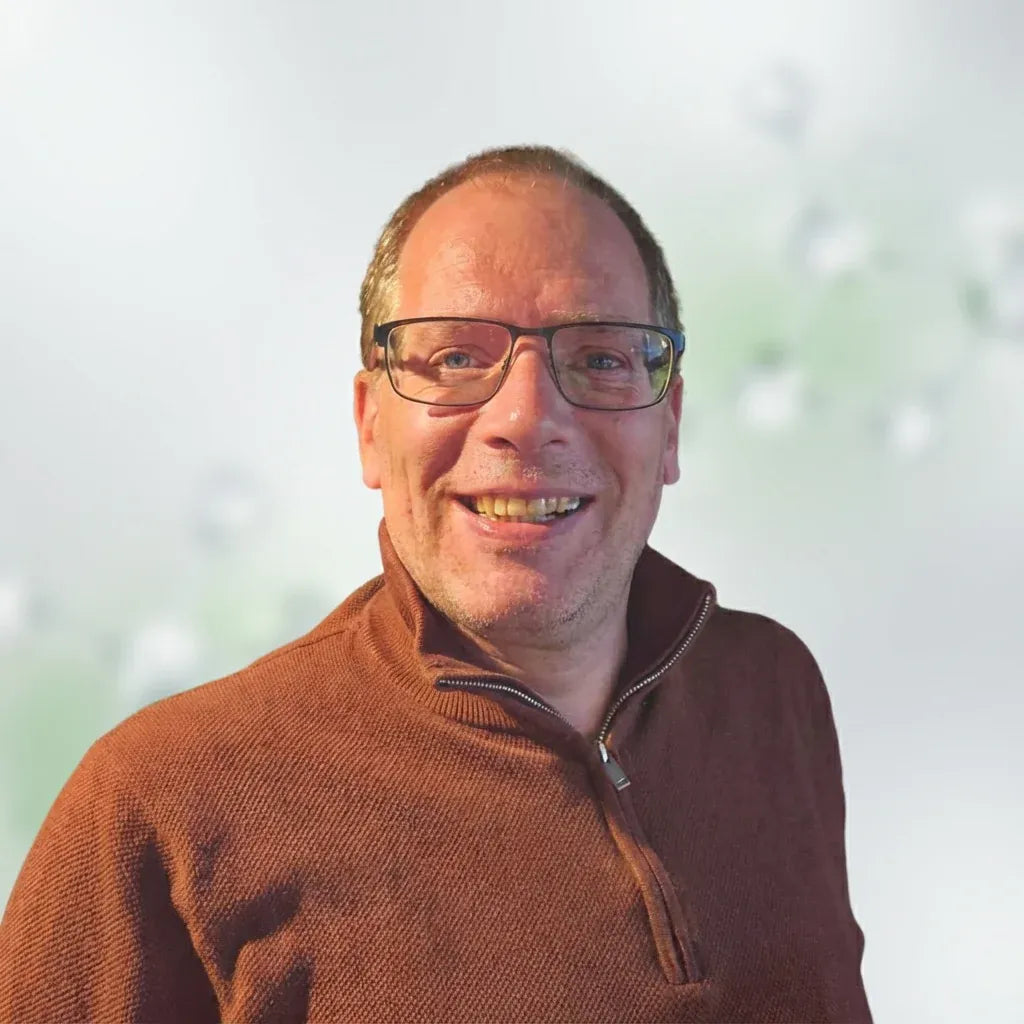 A smiling man with glasses wearing a brown quarter-zip pullover sweater, photographed against a soft, light, blurred background.