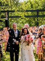 A bride in a white lace wedding gown and floral crown walks down an outdoor aisle with guests showering her with pink flower petals. An older man in a black suit raises his hand in celebration beneath a dark green wooden arch, surrounded by joyful guests and lush green trees.