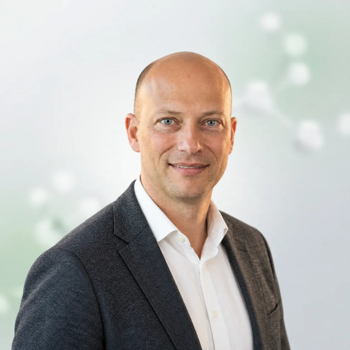 A professional headshot of a smiling bald man with blue eyes, wearing a dark gray blazer over a white collared shirt, set against a soft, light green and white blurred background.