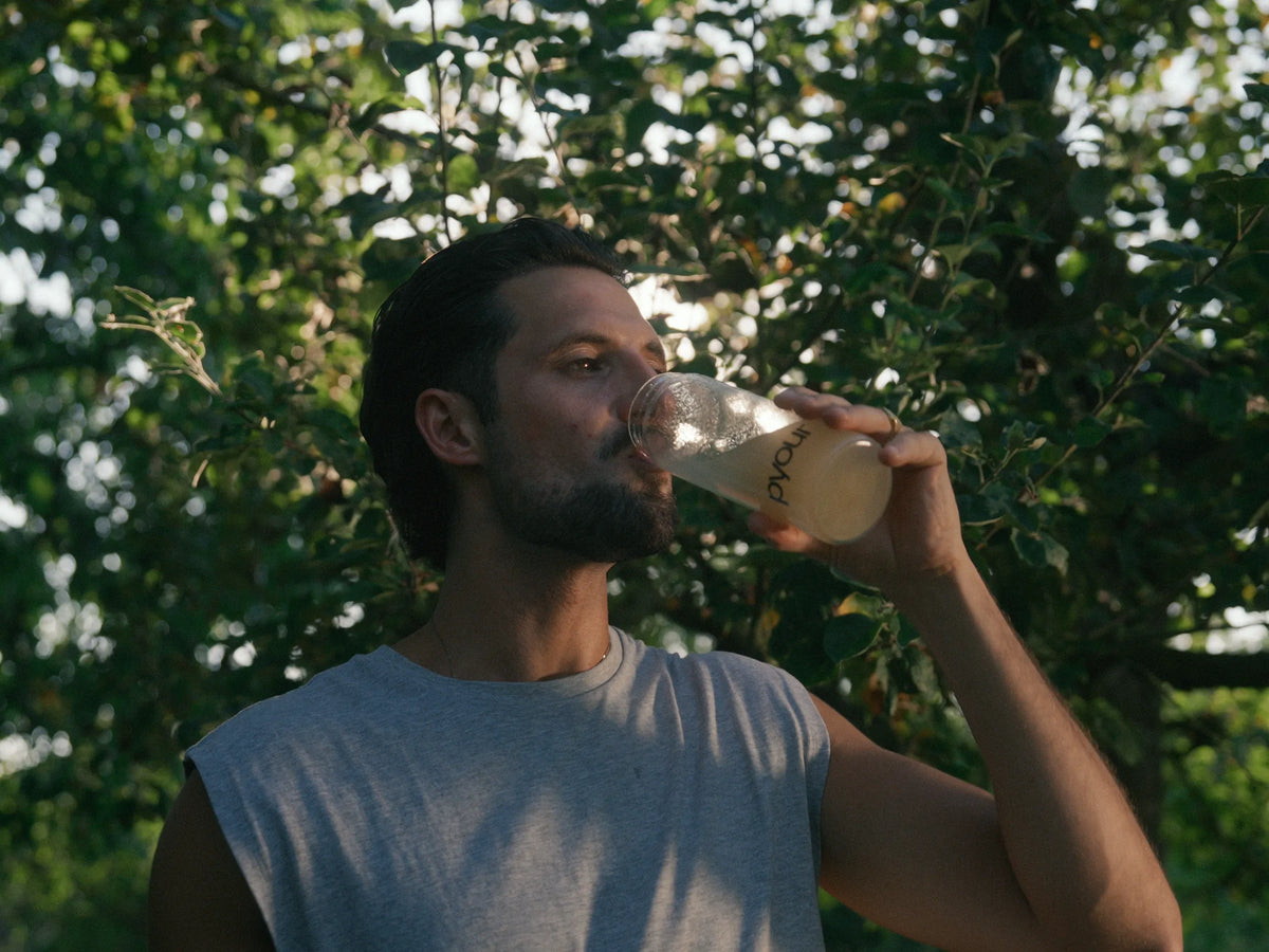 Man drinking Pyour Synergii from a branded glass bottle outdoors, surrounded by green foliage.
