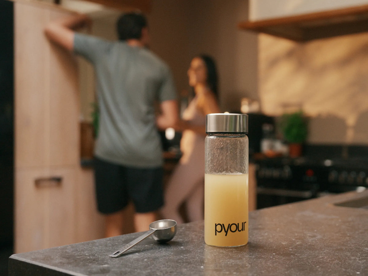 Pyour glass bottle filled with a Synergii drink beside a measuring scoop on a kitchen counter, with a man and woman in the background.