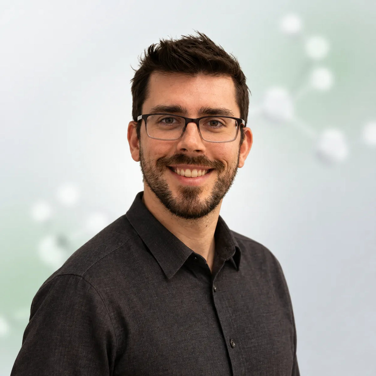 A smiling man with short dark hair, a beard, and glasses wearing a dark charcoal button-up shirt, photographed against a soft light gray and pale green blurred background.