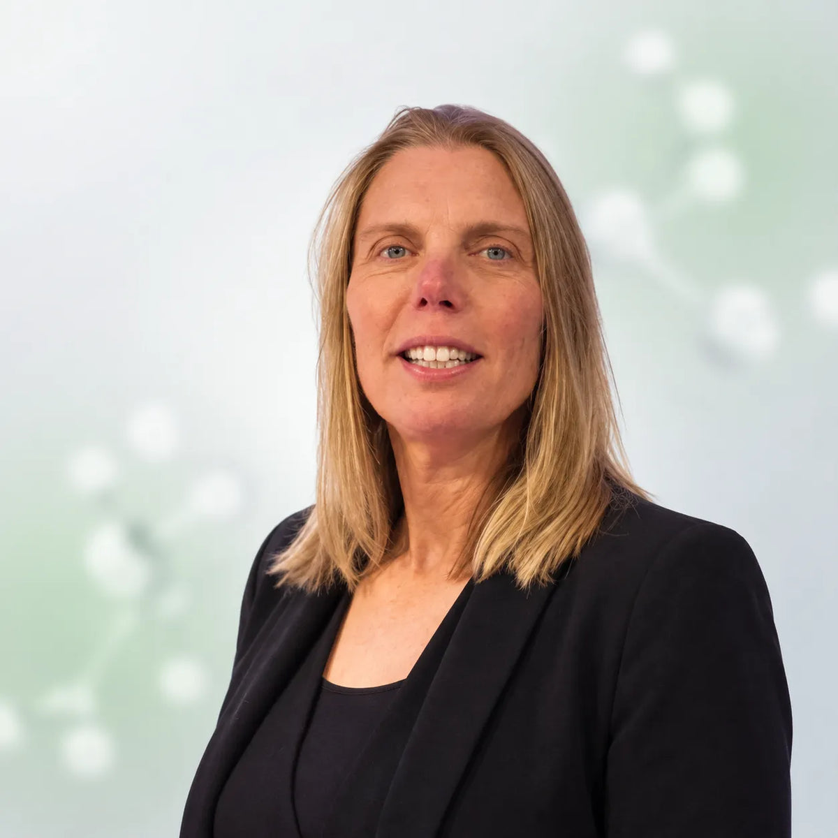 A professional portrait of a smiling blonde woman with blue eyes, wearing a black blazer over a black top, set against a soft, light green and white abstract background.