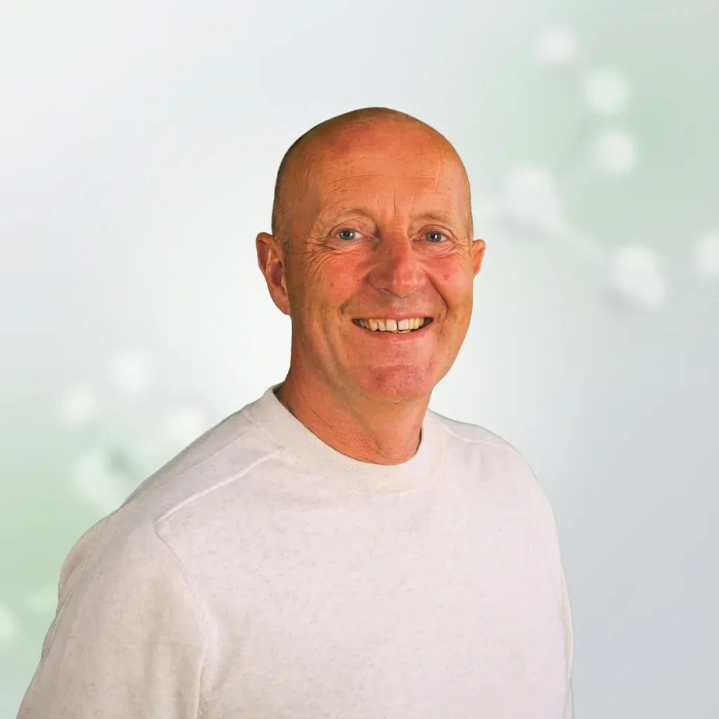 A smiling bald man with blue eyes wearing a light gray crewneck sweatshirt, set against a soft, light green and white bokeh background.